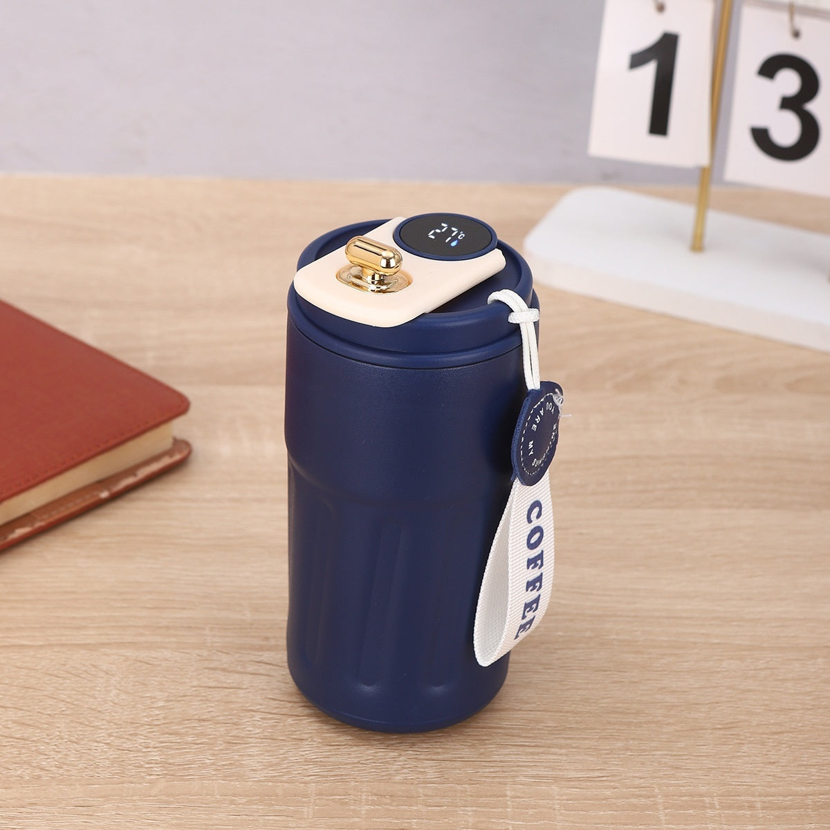 A blue coffee tumbler with a white and gold top, a temperature display, and a white "COFFEE" tag, sitting on a wooden surface next to a brown book and a calendar.
