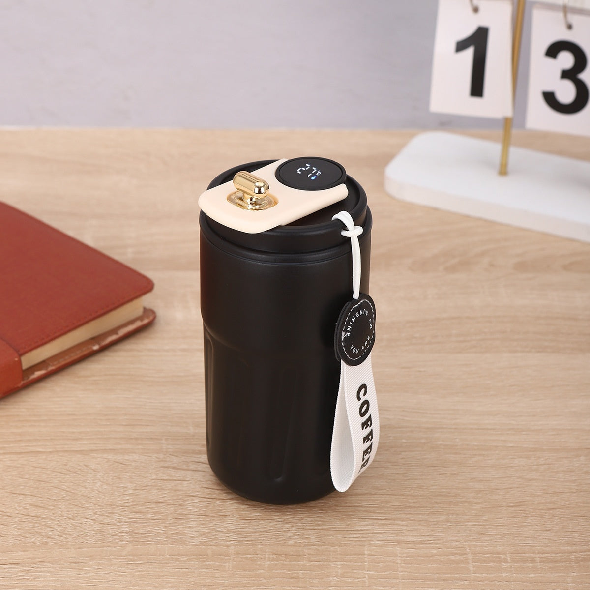 A black travel mug with a digital thermometer lid and a "COFFEE" strap sits on a wooden surface. Beside it is a closed notebook and a calendar.