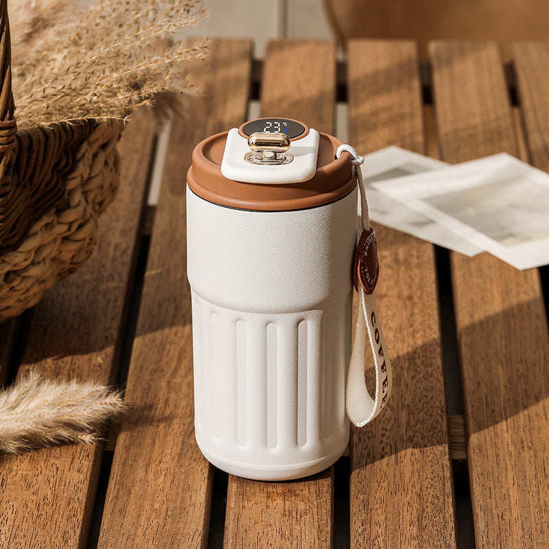 A white thermal coffee mug with brown accents sits on a wooden table, next to a woven basket and some cards.