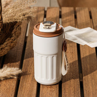 A white thermal coffee mug with brown accents sits on a wooden table, next to a woven basket and some cards.