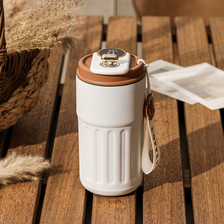 A white thermal coffee mug with brown accents sits on a wooden table, next to a woven basket and some cards.
